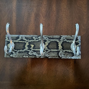 May include: A decorative wall-mounted coat rack with three silver hooks. The rack features a rectangular base covered in a snakeskin pattern in shades of brown, tan, and black. The hooks are curved and made of a shiny metal.