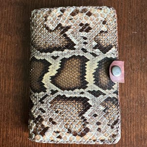 May include: A rectangular wallet featuring a textured snakeskin pattern in shades of brown, tan, and cream. The wallet is secured with a small, dark brown leather tab that includes a single silver-toned metal snap button closure on the right side.