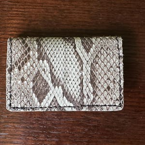 May include: A rectangular card holder with a snakeskin pattern in shades of brown, gray, and white. The edges are neatly stitched with black thread. The accessory is made of a textured material.