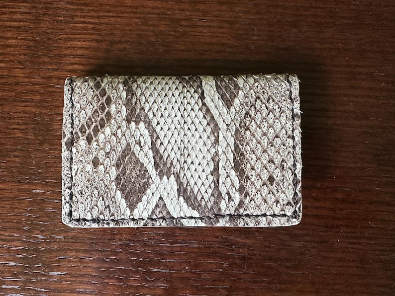 May include: A rectangular card holder with a snakeskin pattern in shades of brown, gray, and white. The edges are neatly stitched with black thread. The accessory is made of a textured material.