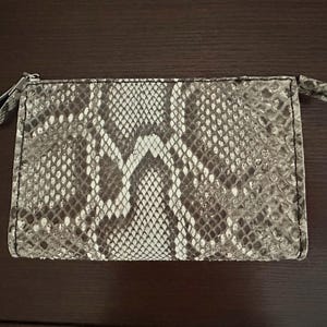 May include: A rectangular, zippered pouch with a snakeskin pattern in shades of gray, brown, and white. The pouch has a small loop on each end and a zipper closure. The overall design is sleek and stylish.
