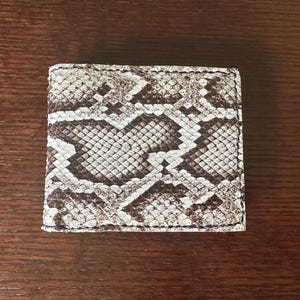 May include: A bi-fold wallet with a snakeskin pattern in shades of brown, tan, and white. The wallet is rectangular and appears to be made of leather. The edges are stitched with black thread. The wallet is closed and sits on a wooden surface.
