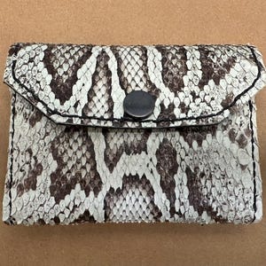May include: A small white and brown snakeskin wallet with a black button closure.
