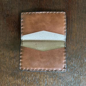 K&ouml;nnte beinhalten: Handgefertigter Lederkartenhalter, offen, um das Innere zu zeigen. Die Brieftasche ist braun mit einem hellbraunen Innenraum und einer kontrastierenden beigefarbenen Tasche. Die Kanten sind mit dunklerem Faden gen&auml;ht. Der Kartenhalter ist rechteckig.