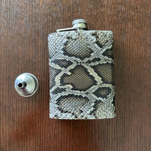 May include: A rectangular stainless steel flask with a snakeskin pattern. The flask has a silver cap and a hinged closure. A small silver funnel sits to the left of the flask. The flask is approximately 4 inches tall.
