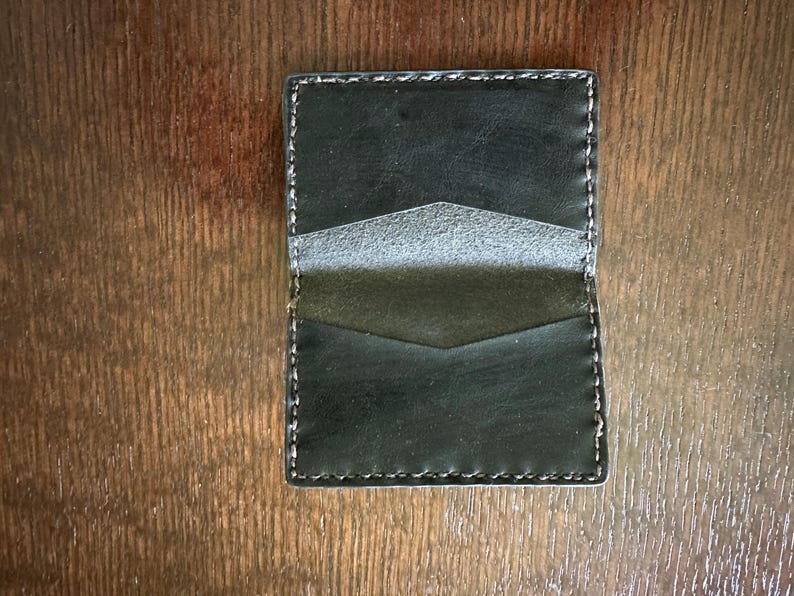 K&ouml;nnte beinhalten: Ein schwarzer Lederkartenhalter mit wei&szlig;en N&auml;hten. Das Portemonnaie hat drei F&auml;cher mit einem kontrastierenden grauen Innenraum. Der Kartenhalter ist rechteckig und liegt auf einer Holzoberfl&auml;che.