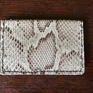 May include: A rectangular card holder made of snakeskin. The card holder features a pattern of white, brown, and black scales. The edges are stitched with black thread. The card holder is a neutral color and is likely used to store credit cards or business cards.