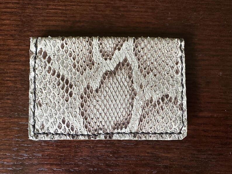 May include: A rectangular card holder made of snakeskin. The card holder features a pattern of white, brown, and black scales. The edges are stitched with black thread. The card holder is a neutral color and is likely used to store credit cards or business cards.