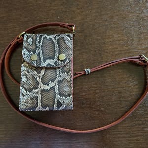 May include: A brown and tan snakeskin-patterned leather phone case with a matching brown leather strap. The case has a flap closure with two brass-colored button snaps. The strap is adjustable and appears to be made of two thin leather cords.