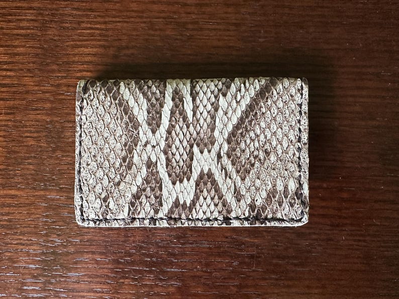 May include: A rectangular card holder with a snakeskin pattern in shades of brown, tan, and white. The wallet is made of a textured material and has a stitched border. The accessory is placed on a wooden surface.