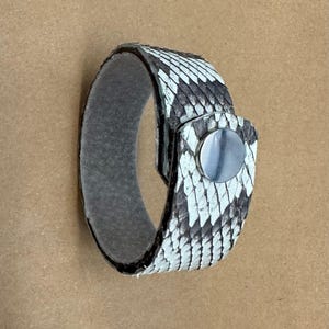 May include: A snakeskin bracelet with a black and white pattern. The bracelet has a silver-colored clasp and a gray interior. The bracelet is shown against a brown background.
