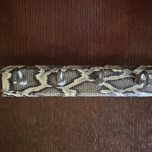 May include: A key holder with a snakeskin pattern. The rectangular holder has four silver-colored hooks. The snakeskin pattern is a mix of tan, brown, and black. The key holder is mounted on a dark brown wooden surface.