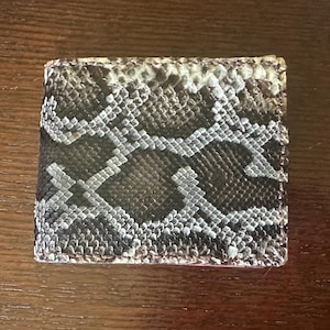 May include: A square-shaped bi-fold wallet featuring a textured snakeskin pattern in shades of dark brown, black, and off-white. The material displays a distinct scale-like surface with a repeating geometric design across the entire exterior surface.