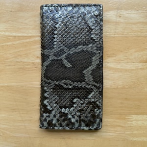 May include: A rectangular wallet with a snakeskin pattern in shades of gray and black. The wallet is made of a textured material and has a dark border. The wallet is displayed on a light brown wooden surface.