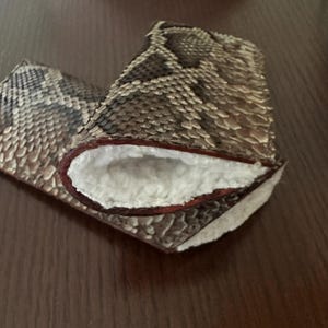 May include: A pair of gloves with a snakeskin pattern in shades of brown, tan, and gray. The interior is lined with white, fluffy material, and the edges are trimmed with a reddish-brown color.