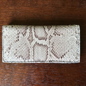 May include: A rectangular wallet featuring a textured snakeskin pattern in shades of beige, cream, and brown. The accessory has a folded design with visible stitching along the edges, showcasing a detailed scale texture across the entire surface.