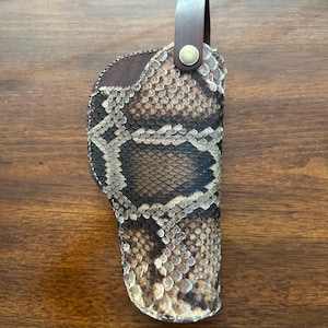 May include: A brown and tan snakeskin-patterned holster with a leather loop and brass button closure. The holster is designed to hold a firearm and is made of a durable material.