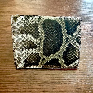 May include: A rectangular wallet with a snakeskin pattern in shades of gray, black, and white. The wallet is made of a textured material and appears to be folded. The wallet is sitting on a wooden surface.