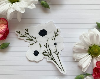May include: A white sticker featuring a bouquet of anemone flowers with black centers and green stems and leaves. The sticker is surrounded by white daisies and red carnations on a lined paper background.