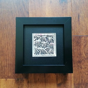 May include: A black framed artwork featuring a silver cutout design of waves. The artwork is signed "November 11" and "10cm x 10cm".