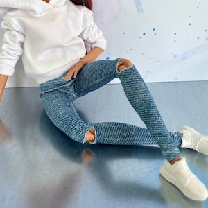May include: A doll wearing a white hooded sweatshirt and blue jeans with distressed details. The doll has long red hair and is sitting on a silver surface.