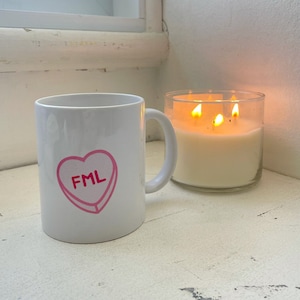 Puede incluir: Taza de cerámica blanca con un diseño de corazón rosa que dice "FML". La taza está sobre una superficie blanca junto a una vela de vidrio transparente con tres mechas encendidas.