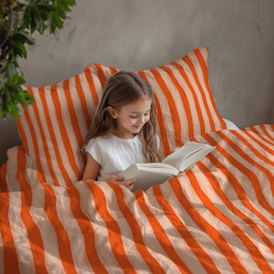 May include: A child reading a book in bed, surrounded by orange and white striped bedding. The duvet cover and pillowcases have the same pattern. The person is wearing a white top.