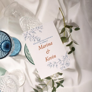 Op de afbeelding: Een witte huwelijksuitnodiging met blauwe bloemenmotieven en de tekst "With heart full of joy we invite you to the wedding of Marina & Kevin".