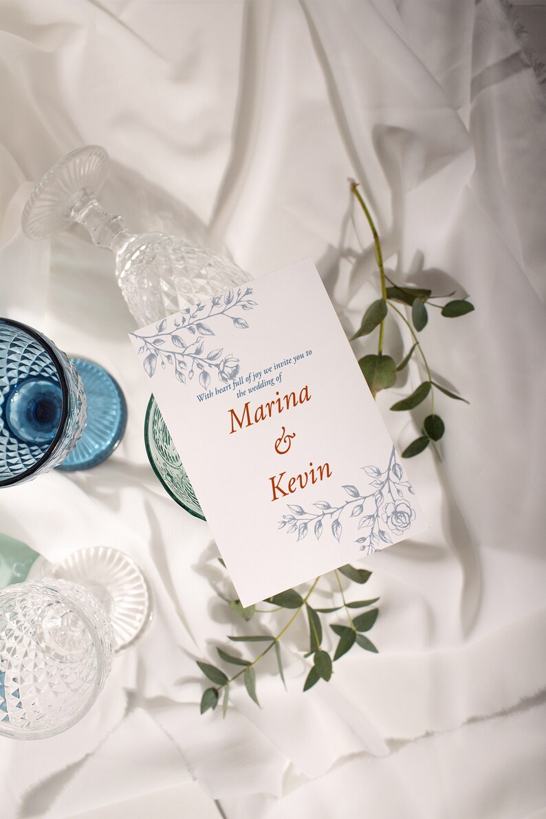 Op de afbeelding: Een witte huwelijksuitnodiging met blauwe bloemenmotieven en de tekst "With heart full of joy we invite you to the wedding of Marina & Kevin".