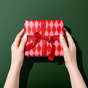 Op de afbeelding: Een ingepakte geschenkdoos met een rode strik, vastgehouden door twee handen. Het cadeaupapier heeft een roze en rood ruitpatroon op een groene achtergrond.