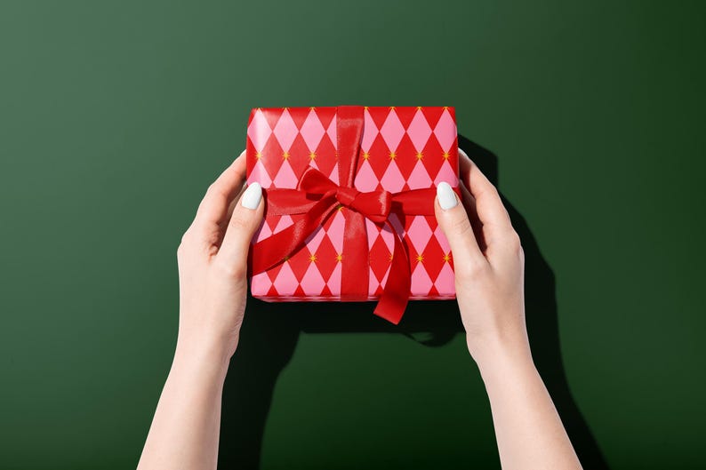 Op de afbeelding: Een ingepakte geschenkdoos met een rode strik, vastgehouden door twee handen. Het cadeaupapier heeft een roze en rood ruitpatroon op een groene achtergrond.