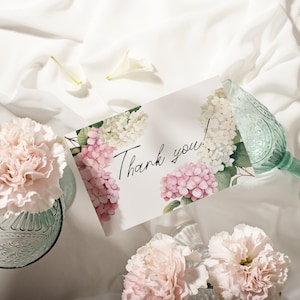 May include: A white card with pink and white hydrangeas printed on it. The card says "Thank you!" in black cursive writing. The card is on a white fabric surface with two clear glass vases with pink carnations.
