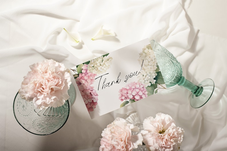 May include: A white card with pink and white hydrangeas printed on it. The card says "Thank you!" in black cursive writing. The card is on a white fabric surface with two clear glass vases with pink carnations.