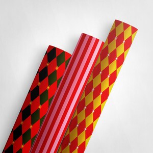 Op de afbeelding: Drie rollen decoratief cadeaupapier. E&eacute;n rol heeft een rood-zwart ruitpatroon met gouden steraccenten. Een andere heeft rood-roze strepen. De derde rol heeft een geel-rood ruitpatroon met witte steraccenten.