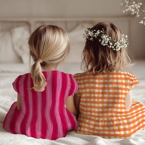 May include: Two young children sit side-by-side on a white surface, viewed from the back. One wears a pink and red striped dress, the other an orange and yellow checkered dress and a floral crown. The scene is soft and light.