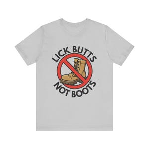 May include: Light gray t-shirt with a graphic design. The design features a brown boot inside a red circle with a line through it, and the words "LICK BUTTS" above and "NOT BOOTS" below in black text.