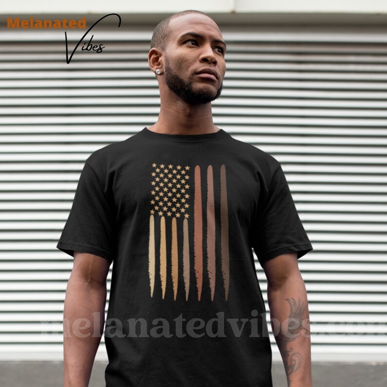 Op de afbeelding: Een zwart T-shirt met een afbeelding van een Amerikaanse vlag met bruine strepen die verschillende huidtinten voorstellen. De tekst "Melinated Vibes" staat in de linkerbovenhoek van de afbeelding.