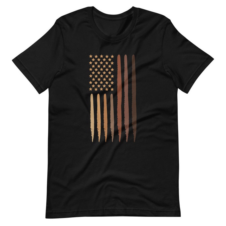 Op de afbeelding: Zwart T-shirt met een afbeelding van een Amerikaanse vlag met strepen in verschillende bruintinten die huidtinten voorstellen.