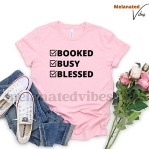 K&ouml;nnte beinhalten: Ein pinkfarbenes T-Shirt mit einer schwarzen Grafik, auf der "BOOKED BUSY BLESSED" mit H&auml;kchen neben jedem Wort steht.