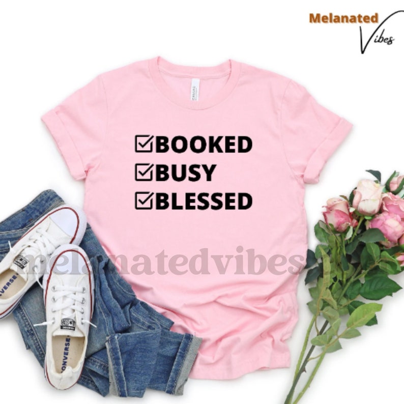 K&ouml;nnte beinhalten: Ein pinkfarbenes T-Shirt mit einer schwarzen Grafik, auf der "BOOKED BUSY BLESSED" mit H&auml;kchen neben jedem Wort steht.