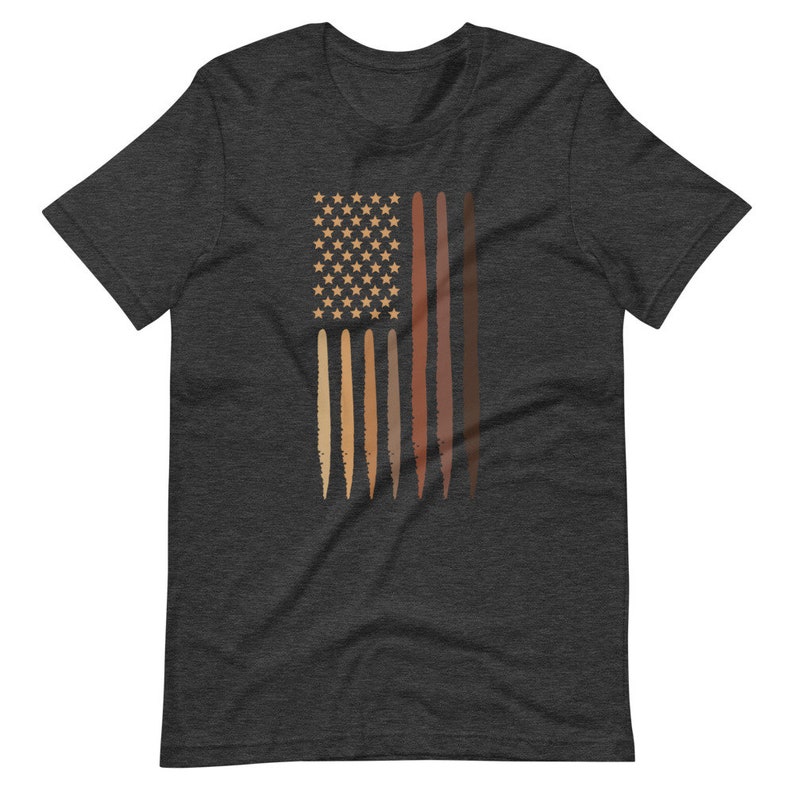 Op de afbeelding: Een donkergrijs t-shirt met een afbeelding van een Amerikaanse vlag met strepen die verschillende huidtinten voorstellen. De sterren zijn wit.