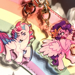 Könnte beinhalten: Zwei bunte Acryl-Schlüsselanhänger mit Cartoon-Pferdefiguren mit Flügeln. Der erste Schlüsselanhänger zeigt ein rosa, blaues und violettes Pony mit einem rosa und blauen Flügel. Der zweite Schlüsselanhänger zeigt ein violettes Pony mit einem rosa Flügel und einer rosa Blume im Haar. Beide Ponys halten einen kleinen Gegenstand in ihren Hufen.