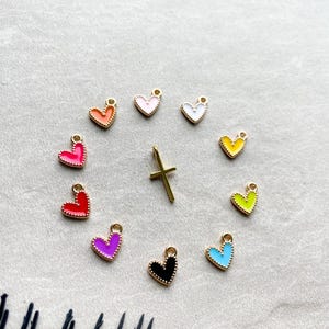 Könnte beinhalten: Eine Sammlung bunter herzförmiger Anhänger und ein goldener Kreuzanhänger. Die Herzanhänger sind in verschiedenen Farben erhältlich, darunter Rosa, Orange, Rot, Lila, Schwarz, Blau, Gelb und Grün. Jeder Anhänger hat einen goldenen Rand und eine kleine Schlaufe.