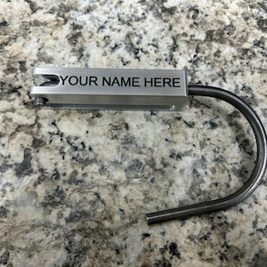 May include: A silver-colored metal key tag with the text "YOUR NAME HERE" engraved on it. The key tag has a curved, dark metal loop attached. The background is a speckled, light-colored surface.