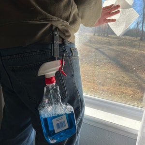 Może przedstawiać: Niebieska butelka z rozpylaczem z czerwonym spustem i białą etykietą z napisem "64 oz. FL. OZ. Multi-Surface Cleaner" jest przymocowana do metalowego haka. Hak jest przymocowany do pętli paska na niebieskich dżinsach.