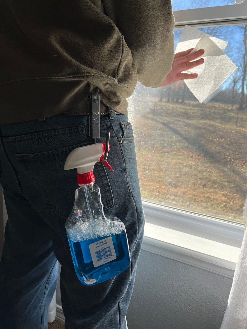 Może przedstawiać: Niebieska butelka z rozpylaczem z czerwonym spustem i białą etykietą z napisem "64 oz. FL. OZ. Multi-Surface Cleaner" jest przymocowana do metalowego haka. Hak jest przymocowany do pętli paska na niebieskich dżinsach.