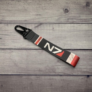 May include: A black fabric keychain strap featuring the white and red N7 logo. The strap includes a black metal lobster clasp at the top and a distressed, weathered design with red and white stripes near the ends of the black fabric band.