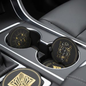 May include: Black car coasters with gold designs and text. One coaster features a sun-like design, another has a floral motif with text, and a third displays a geometric pattern. The coasters are in a car's cup holders.