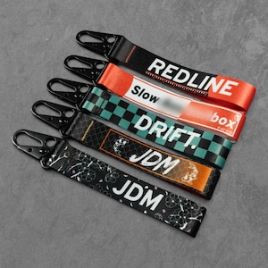 May include: Five fabric keychains with black metal clips. The straps feature various designs: red with white text reading REDLINE, orange and white with Slow box text, a teal and black checkerboard with DRIFT, black with JDM and koi fish, and black with JDM and cherry blossoms.