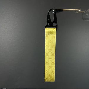 K&ouml;nnte beinhalten: Ein gelbes Stoff-Handgelenkband mit einem sich wiederholenden Chevron-Muster, befestigt an einem schwarzen Metallkarabiner. Das Band besteht aus gewebtem Material mit gen&auml;hten Kanten und h&auml;ngt vertikal an einem silbernen Metallhaken.
